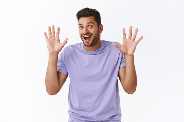 Cheerful enthusiastic modern guy in purple t-shirt have nothing hide, raising hands in surrender or retreatment, smiling joyfully, waving hands in hi, friendly greeting gesture, white wall