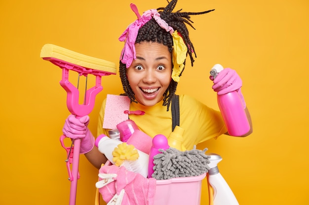 Cheerful ethnic housewife has braids poses with mop and detergent bottle happy to clean