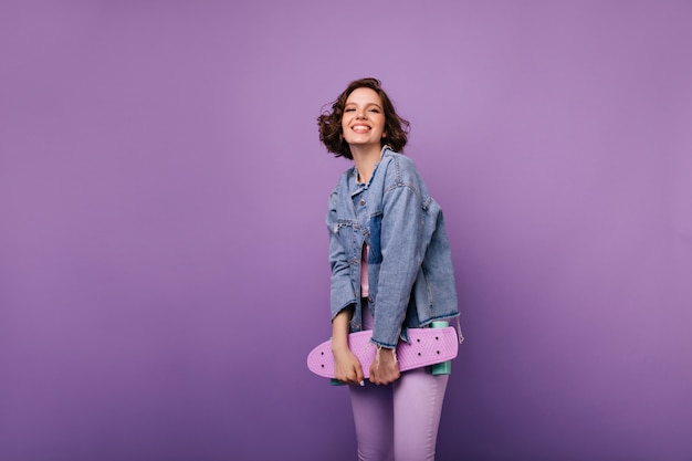 Free photo cheerful european woman in purple pants posing with skateboard. indoor shot of appealing smiling girl with dark wavy hair.