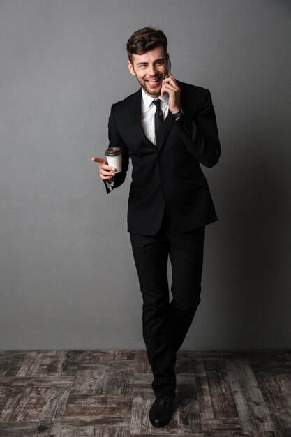 Cheerful excited young business man drinking coffee talking by phone