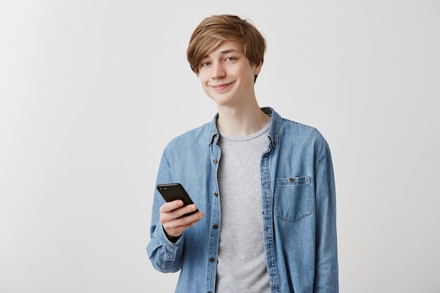 Cheerful fair-haired caucasian male student in danim shirt browsing internet on smartphone, having rest after classes, looking at screen and smiling. Modern technologies and communication