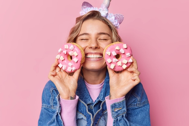 Free photo cheerful fair haired young european woman keeps eyes closed smiles toothily holds two donuts with marshmallow glad to eat favorite dessert dressed in denim jacket isolated over pink background