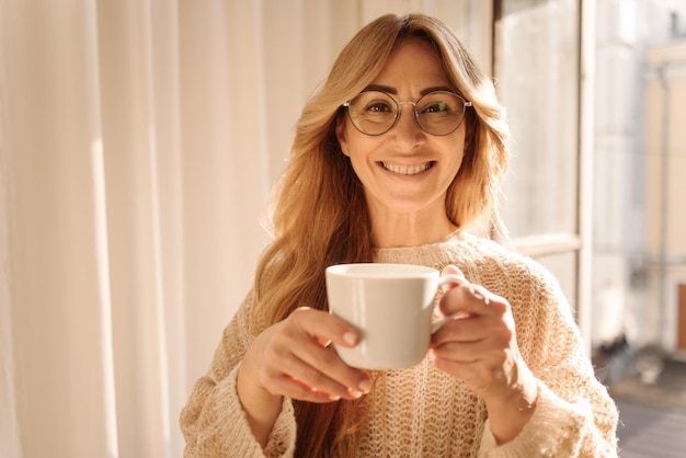 Cheerful fairskinned adult blonde woman in glasses smiling at camera while drinking aromatic coffee while standing by window Leisure relaxation and lifestyle concept