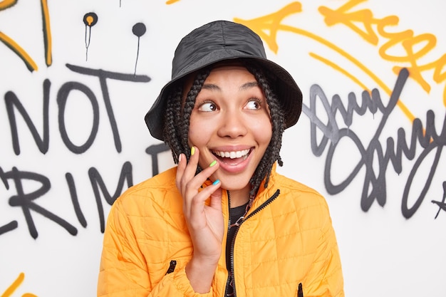 Free photo cheerful fashionable female youngster smiles happily looks aside wears stylish clothes spends free time in urban place poses against colorful graffiti wall