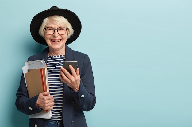 Free photo cheerful female moderator socialices online, holds modern gadget, receives notification, holds papers with notepad, dressed in stylish formal wear, isolated over blue wall, blank space