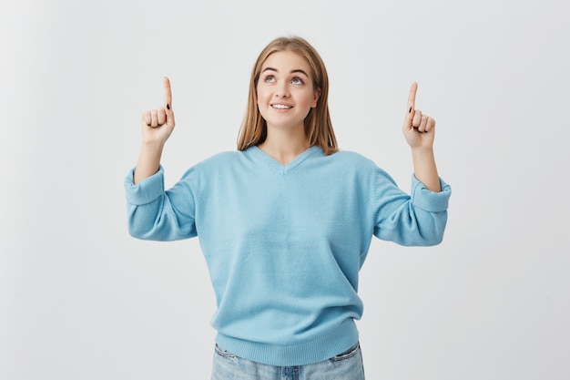 Cheerful female with fair hair, wearing blue sweater and jeans, pointing upwards with her fingers being pleased to see something. Blonde-haired girl joyfully smiling with teeth