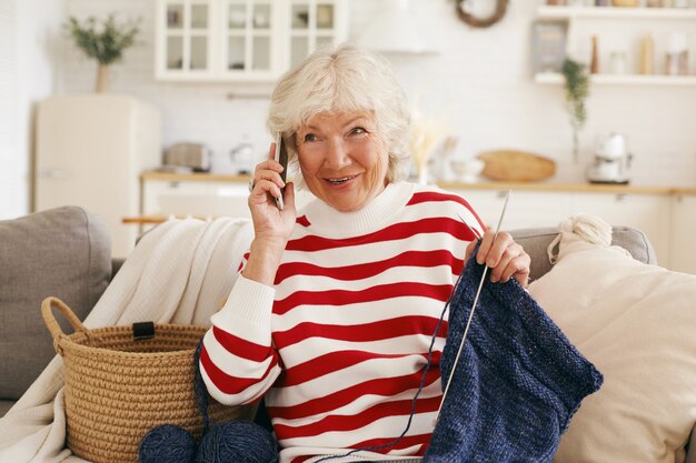 Cheerful friendly looking gray haired senior woman dressed in casual clothes sitting on sofa with needles and yard, having nice phone conversation with her old friend, gossiping, sharing latest news