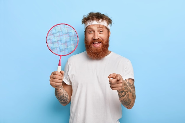 Free Photo cheerful friendly red haired tennis player holds racket while posing against the blue wall