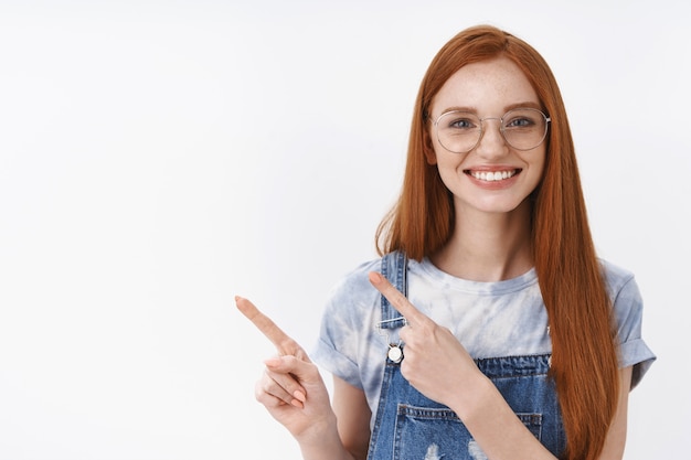 Free photo cheerful friendly sincere redhead girl with blue eyes freckles long red hair pointing left, smiling polite and sociable introduce promo, stand white wall amused joyful, happy help