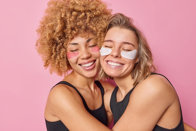 Free photo cheerful friendly women embrace each other with love smile gently apply beauty patches under eyes for skin treatment and rejuvenation dressed in black t shirts isolated over pink background