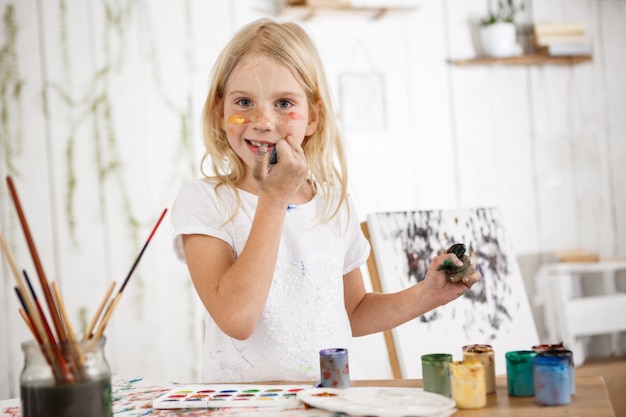 Free photo cheerful and full of joy creative little blonde smiling  and touching her face with dirty hands