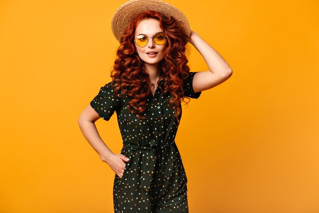 Cheerful ginger girl in sunglasses posing with hand in pocket. Studio shot of refined caucasian lady in straw hat isolated on yellow background.