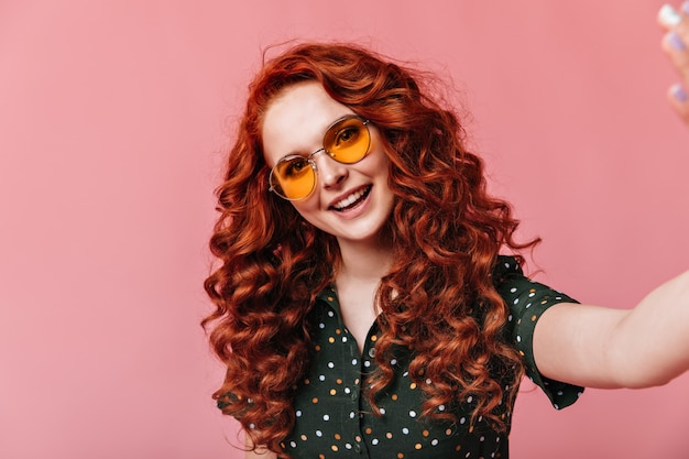 Cheerful ginger woman taking selfie. Studio shot of curious curly girl in sunglasses posing on pink background.