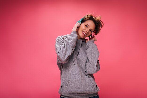 Free Photo cheerful girl in bright headphones posing on isolated background. pretty young woman in grey hoodie listening to music on pink backdrop