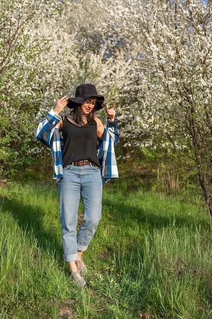 Cheerful girl in a hat among the flowering trees in the spring, in a casual style.