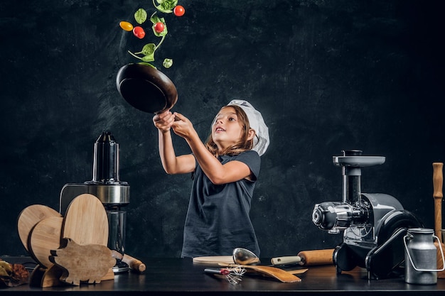 Cheerful girl is tossing vegetables on the pan at dark photo studio.