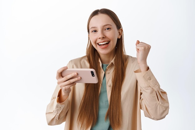 Cheerful girl rejoicing celebrating achievement in mobile app holding horizontal smartphone and make fist pump smiling pleased achieve goal in application white background