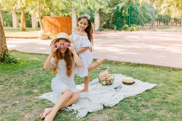 Free photo cheerful girl standing on one leg while her funny mother fooling around with cookies. outdoor portrait of joking long-haired woman enjoying picnic with daughter in vacation.