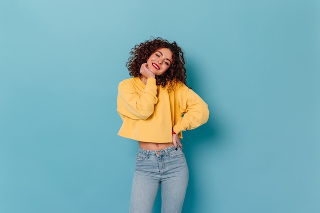 Free Photo cheerful girl with red lips and snow-white smile looks into camera on blue space. portrait of cute curly lady in yellow top with long sleeves and jeans.