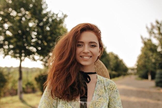 Cheerful girl with wavy ginger hairstyle and black bandage on her neck in summer stylish dress and straw hat smiling and looking at front outdoor