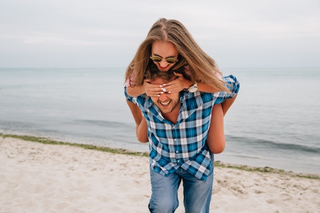 Free photo cheerful guy holding her girlfriend behind the back, while she covering his eyes by hands