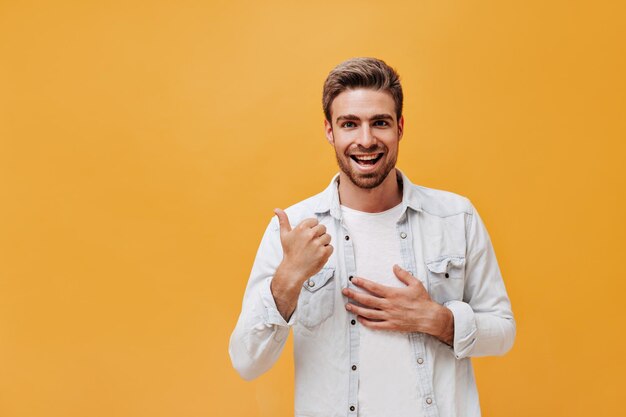 Cheerful guy with beard in white tshirt and denim jacket looking into camera and smiling on orange isolated background