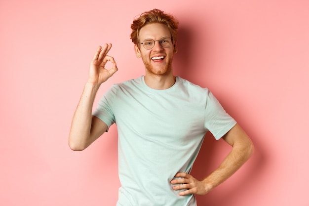 Cheerful guy with red hair and beard, wearing glasses, showing OK sign in approval and saying yes, smiling satisfied, standing over pink background.