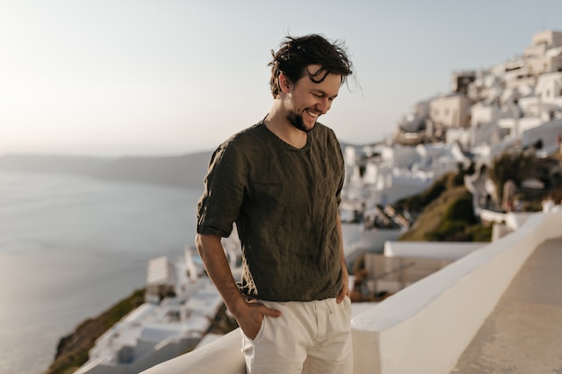 Cheerful happy brunette man in dark-green t-shirt and white shorts smiles sincerely outside