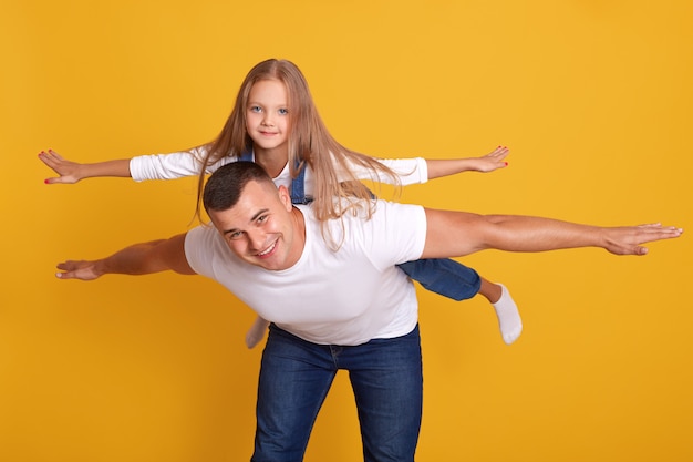 cheerful happy man imagines with his lovely daughter to fly like plane, posing isolated on yellow. shot, happy moments with best father, togetherness, family concept.