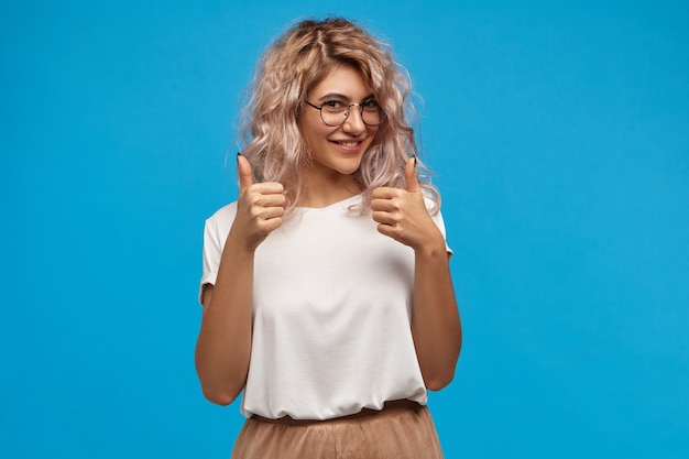 Free Photo cheerful hipster girl in trendy round eyewear making thumb up gesture with both hands and smiling joyfully, showing her support and respect to someone, saying good job, well done