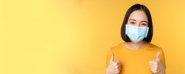 Cheerful korean woman in medical face mask support people during pandemic wear personal protective equipment from covid19 showing thumbs up in approval yellow background