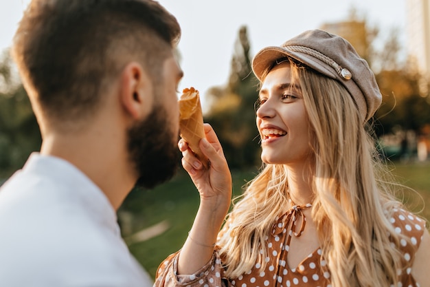 Cheerful lady in stylish hat and polka-dot top laughs,smudging her husbands nose with melon ice cream.