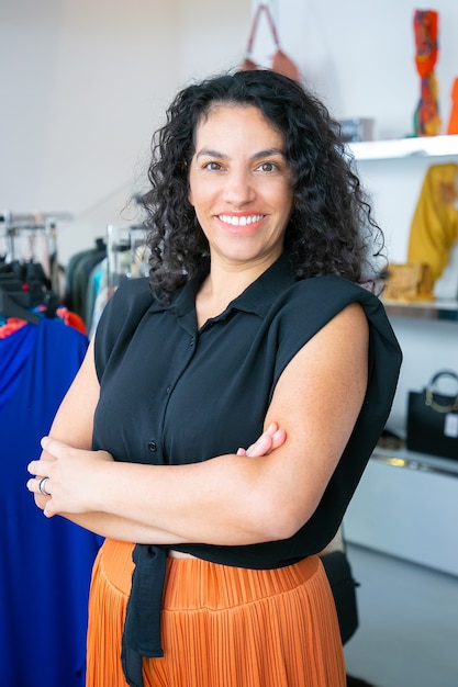 Free Photo cheerful latin black haired woman standing with arms folded near rack with dresses in clothes shop, looking at camera and smiling. boutique customer or shop assistant concept