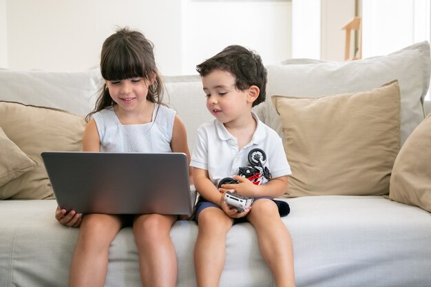 Cheerful little brother and sister sitting on couch at home, using laptop, watching video, cartoon films or funny movie.