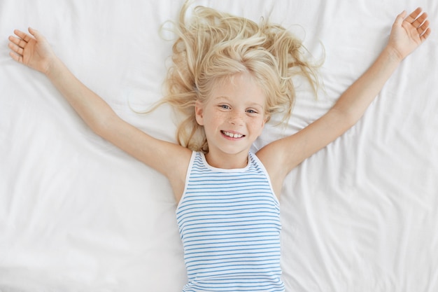 Free photo cheerful little girl with light hair, lying in comfortable bed on white bedclothes, stretching after night sleep, looking with delightful expression. freckled small child relaxing in bed