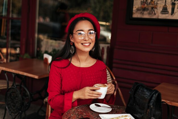 Cheerful longhaired brunette woman in bright beret and red dress smiles widely and sits in beautiful cafe Young Asian lady in eyeglasses holds white coffee cup