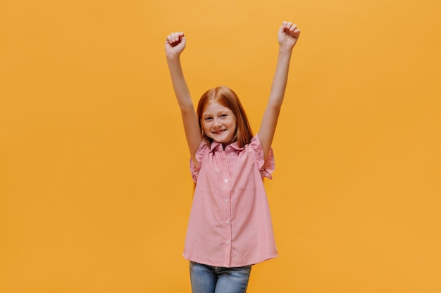 Free photo cheerful longhaired redhead little girl in jeans and striped shirt rises arms and rejoices on isolated orange background