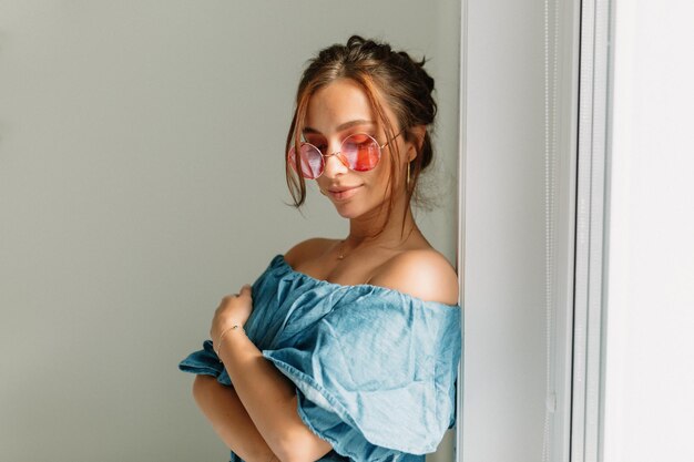 Cheerful lovely woman with dark hair wearing pink round glasses wearing blue tshirt with bare shoulders posing near the window in sunlight