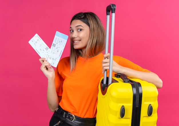 Free photo a cheerful lovely young woman in an orange t-shirt showing plane tickets with yellow suitcase on a pink wall