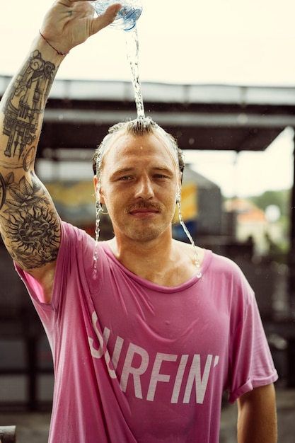 Free photo a cheerful man in a bright t-shirt with a skateboard in a skatepark pours water from a bottle on his head.