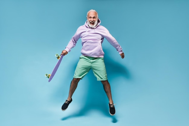 Free Photo cheerful man in hoodie jumping with skateboard on blue background happy bearded guy in purple clothes and shirt jumping