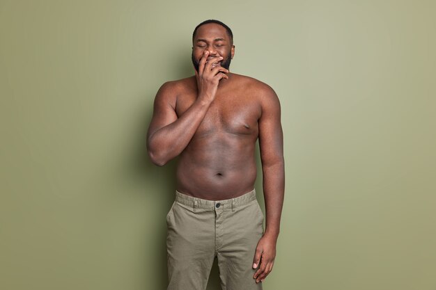 Cheerful man laughs positively covers mouth with hand hears something funny wears trousers shows naked torso has muscular arms poses against khaki studio wall