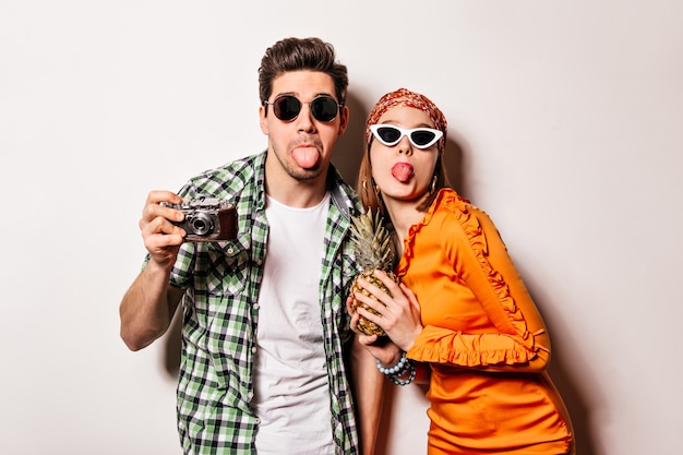 Cheerful man and woman in sunglasses are showing tongues and posing with retro camera and pineapple on isolated space.