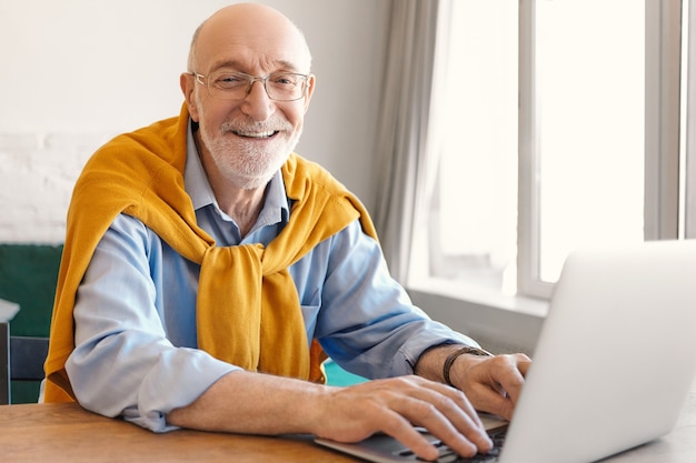 Cheerful mature bearded bald male entrepreneur wearing glasses and sweater over blue formal shirt smiling happily while keyboarding on portable computer, playing video games during lunch break