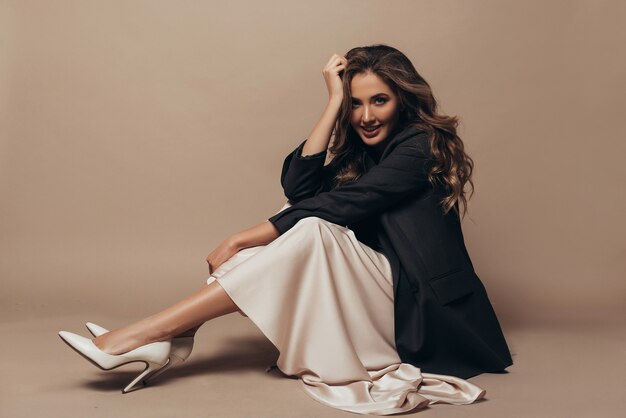 Cheerful model sitting on the floor, wearing modern oversize black jacket and creamy long dress, high heel shoes on her feet. Curly hairstyle and makeup