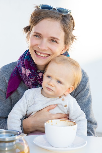 Cheerful mom holding sweet baby in arms while drinking coffee in coffee shop