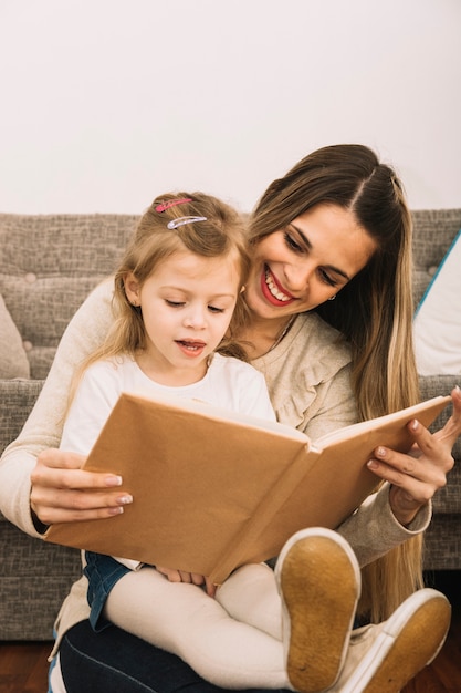 Free photo cheerful mother reading book to daughter near sofa