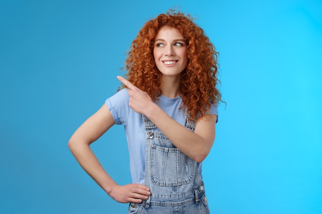 Cheerful motivated charismatic good-looking happy smiling redhead 25s curly-haired woman summer dungarees pointing upper left corner look aside pleased show promo suggest try-out. Copy space