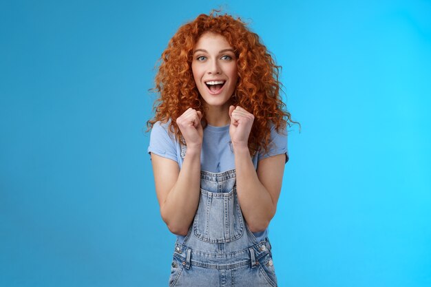 Cheerful optimistic lucky redhead curly-haired attractive woman cheering clench fists joyfully smiling broadly cheering watching game supportive encourage keep up motivated blue background.