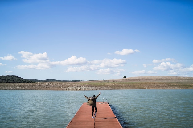 Free photo cheerful person jumping on a dock near the lake under a cloudy sky